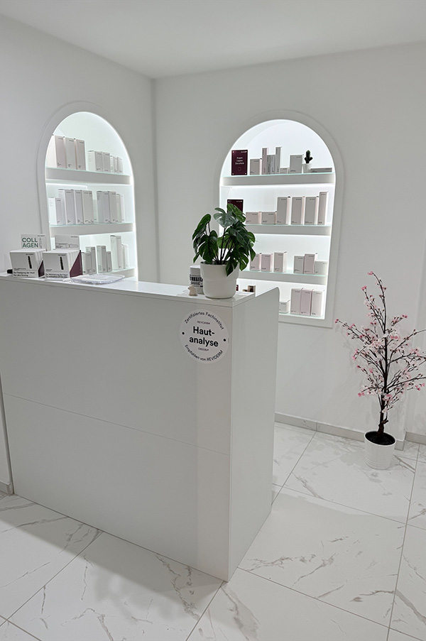 Eine elegante Rezeption einer Hautpflegeklinik mit weißem Tresen, Topfpflanze und übersichtlichen Produktregalen. Die Atmosphäre ist sauber, modern und einladend.