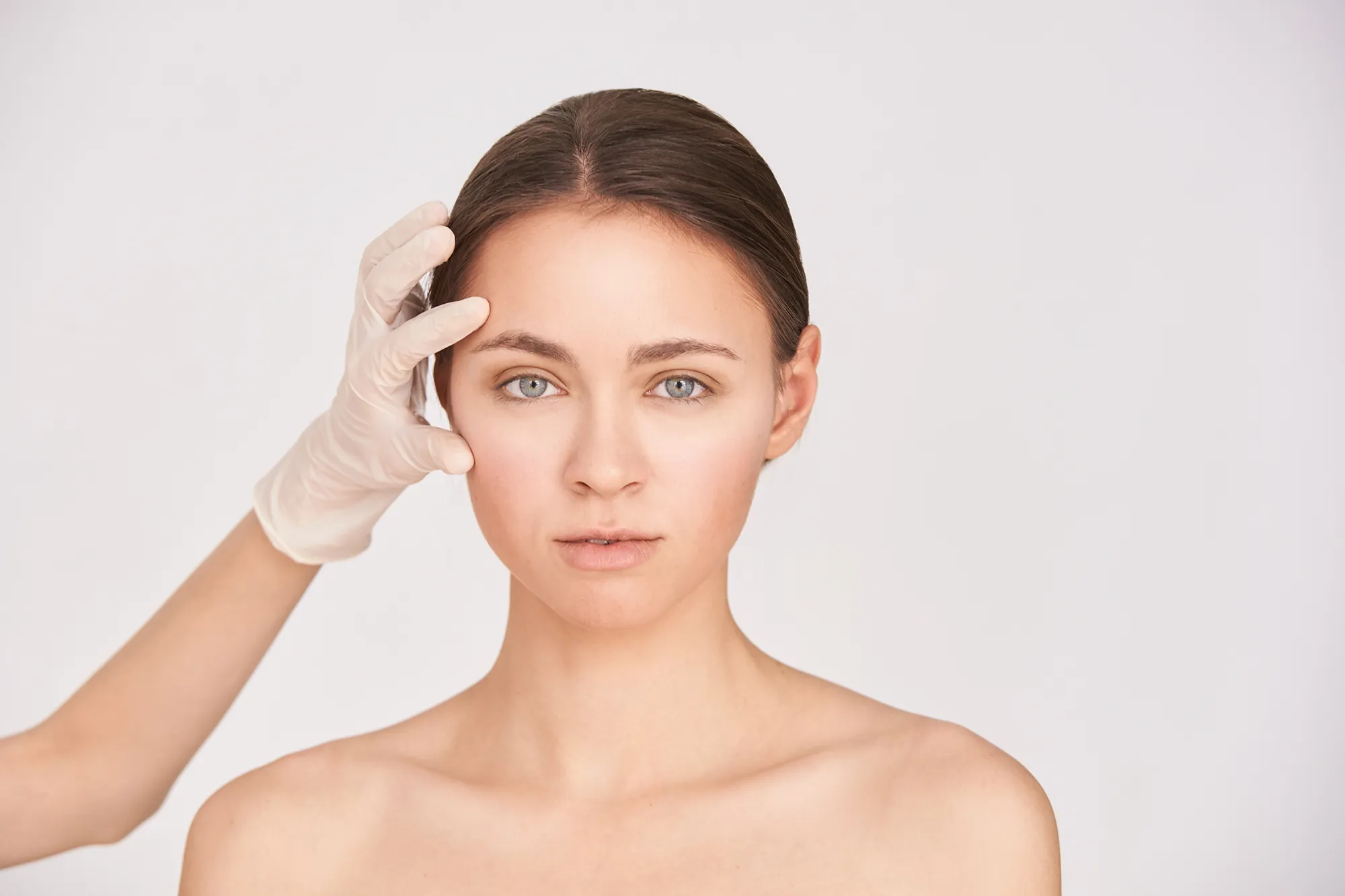 Eine Frau mit neutralem Gesichtsausdruck wird vor einem weißen Hintergrund von einer behandschuhten Hand leicht an der Schläfe berührt, was auf einen klinischen oder Hautpflege-Kontext hindeutet.